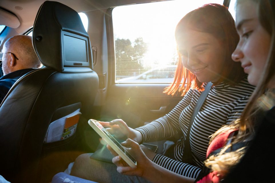Woman sitting beside her sister in a car looking at a phone together while she is on her way to move cities to attend university.